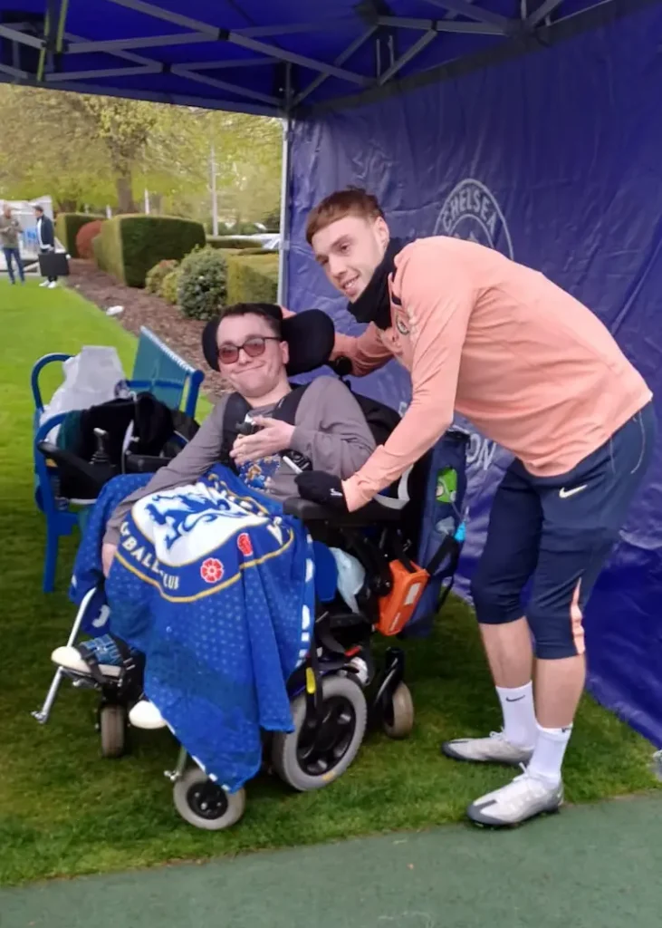 CCS client in a wheelchair, smiling with a Chelsea FC player, under a blue tent with the Chelsea logo visible.