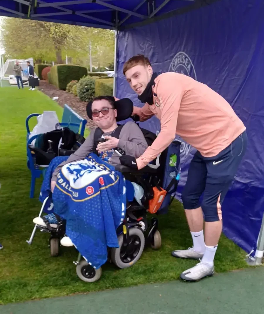 Day Care special outing—celebrating a 21st birthday at Chelsea FC with Continuity of Care Services A young client in a wheelchair smiling alongside the Chelsea FC badge on the pitch during a special day-care outing