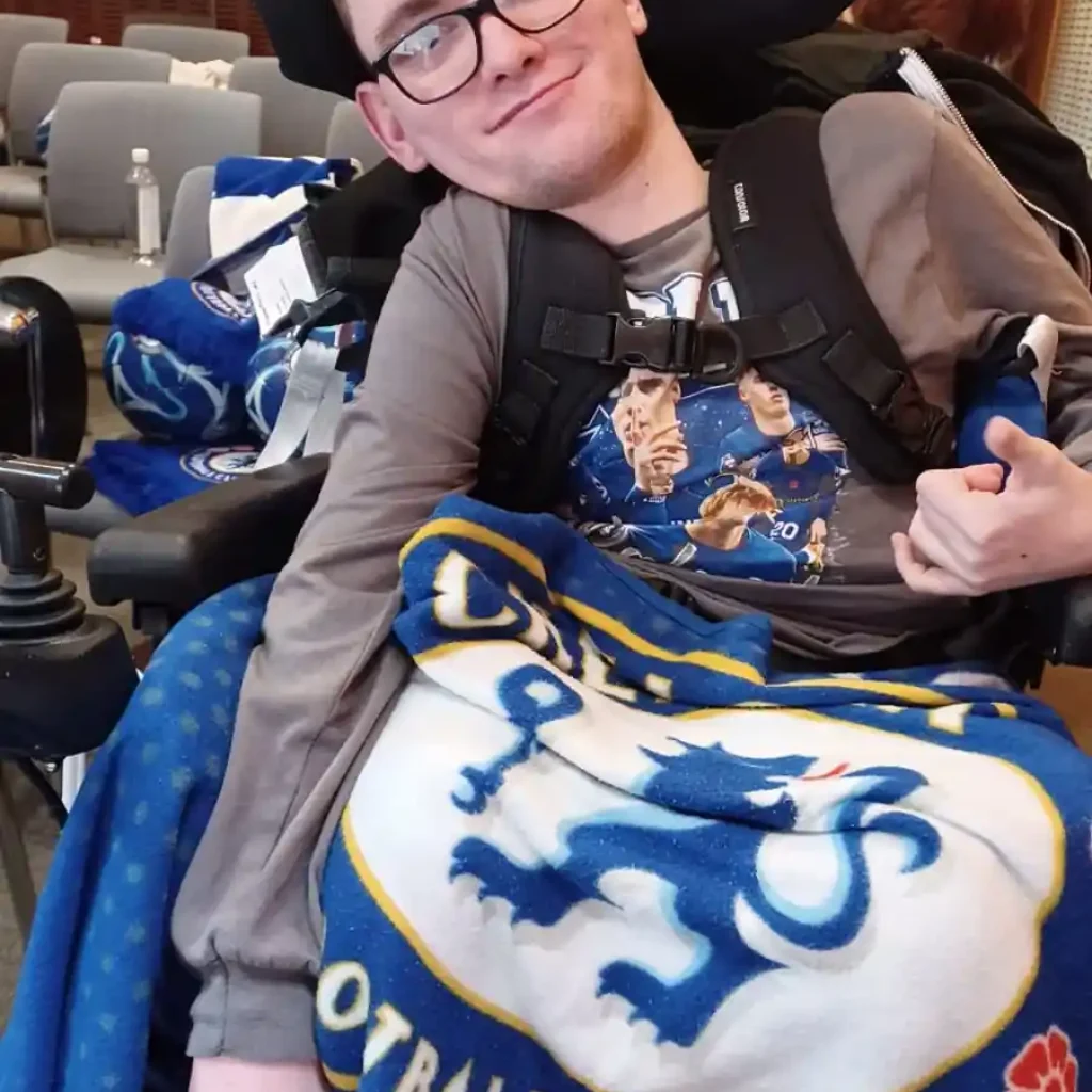 Complex care support—confidence and community for clients in Maidstone & Kent A young man in a power wheelchair wrapped in a Chelsea Football Club blanket, smiling confidently in a community setting.