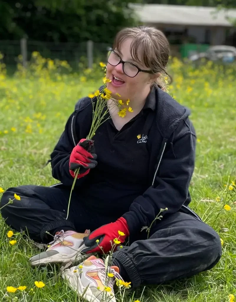 Companionship care, enjoying nature and shared moments in Maidstone & Kent A joyful client seated in a meadow, holding a handful of yellow wildflowers with the support of a friendly carer