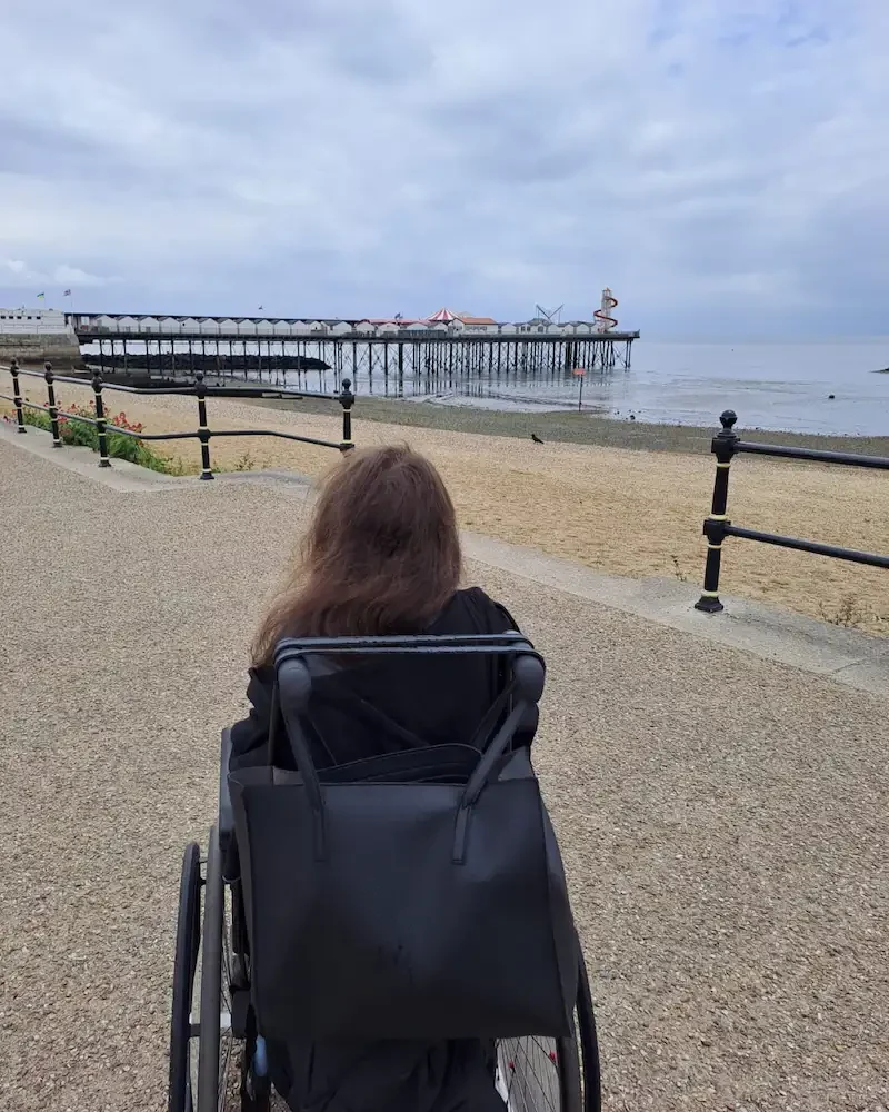 Respite care, refreshing seaside outings for clients in Maidstone & Kent A client in a wheelchair enjoying a peaceful seaside promenade with a pier in the distance