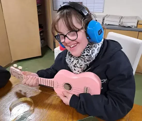 Respite care client enjoying a music session Smiling young woman with glasses and ear defenders playing a pink ukulele